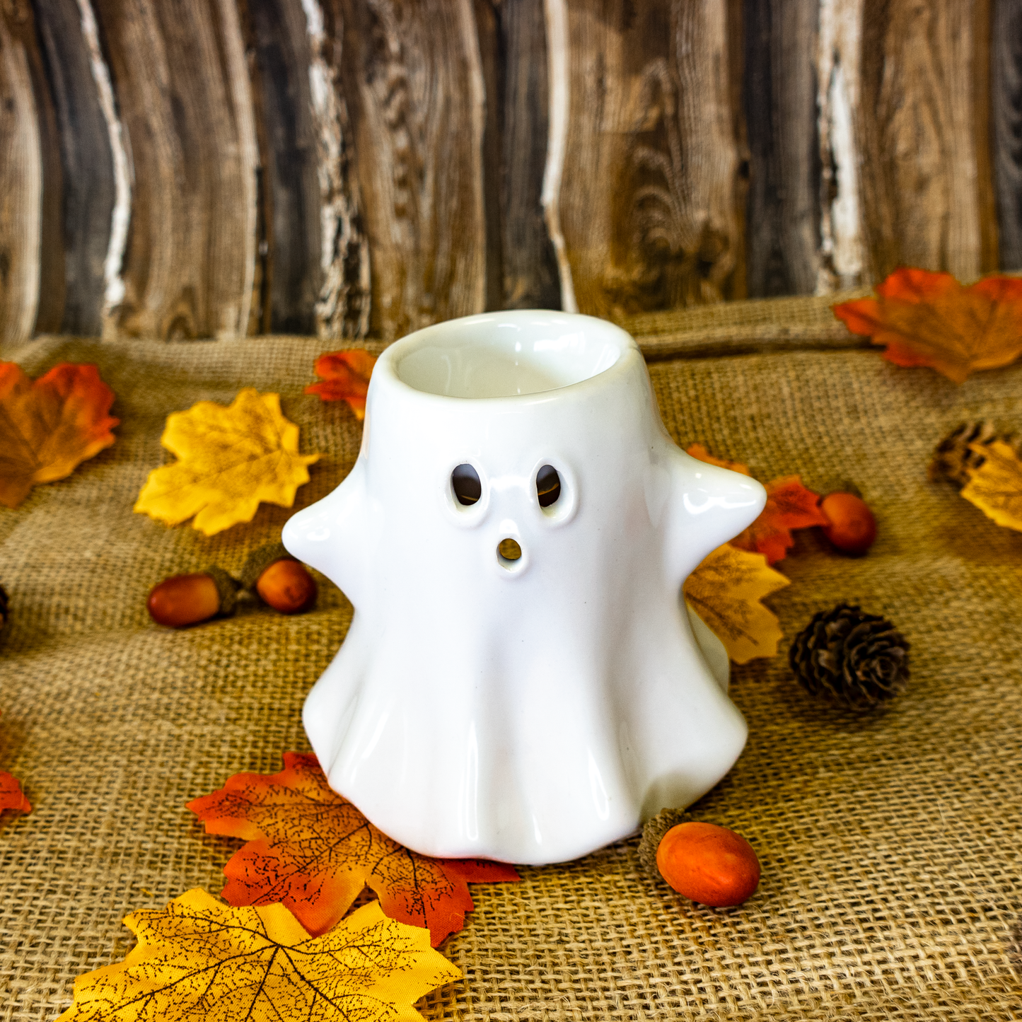 White ghost-shaped candle holder on a burlap surface with autumn leaves and pinecones.
