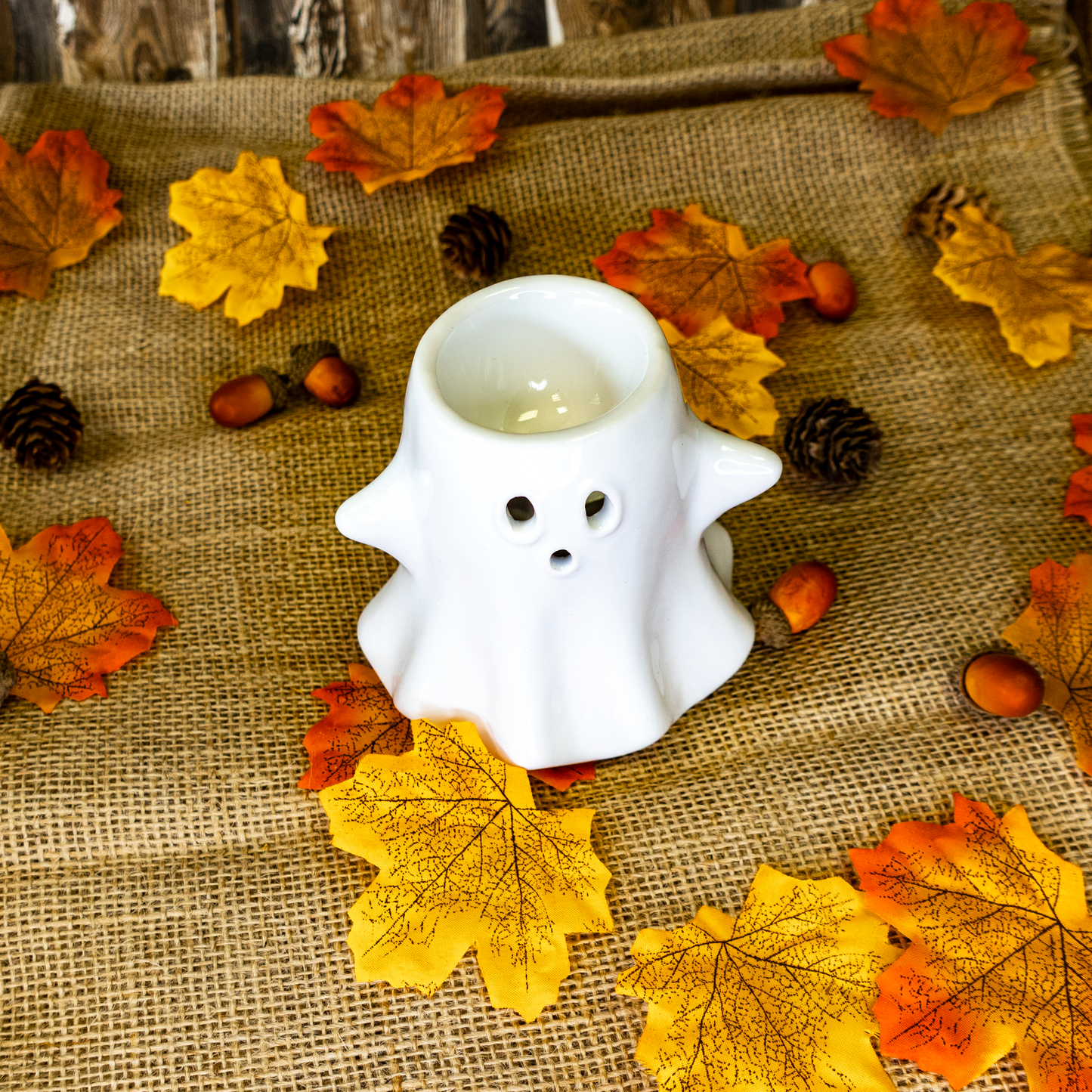 White ghost-shaped candle holder on a burlap surface with autumn leaves and pinecones.