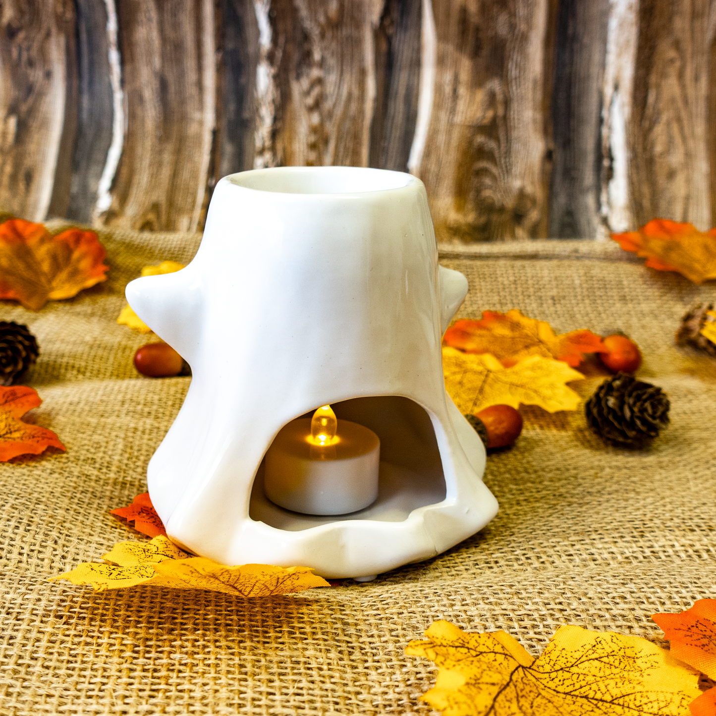 White ceramic wax melter with a lit electric candle inside, surrounded by autumn leaves and pinecones on a rustic wooden background.