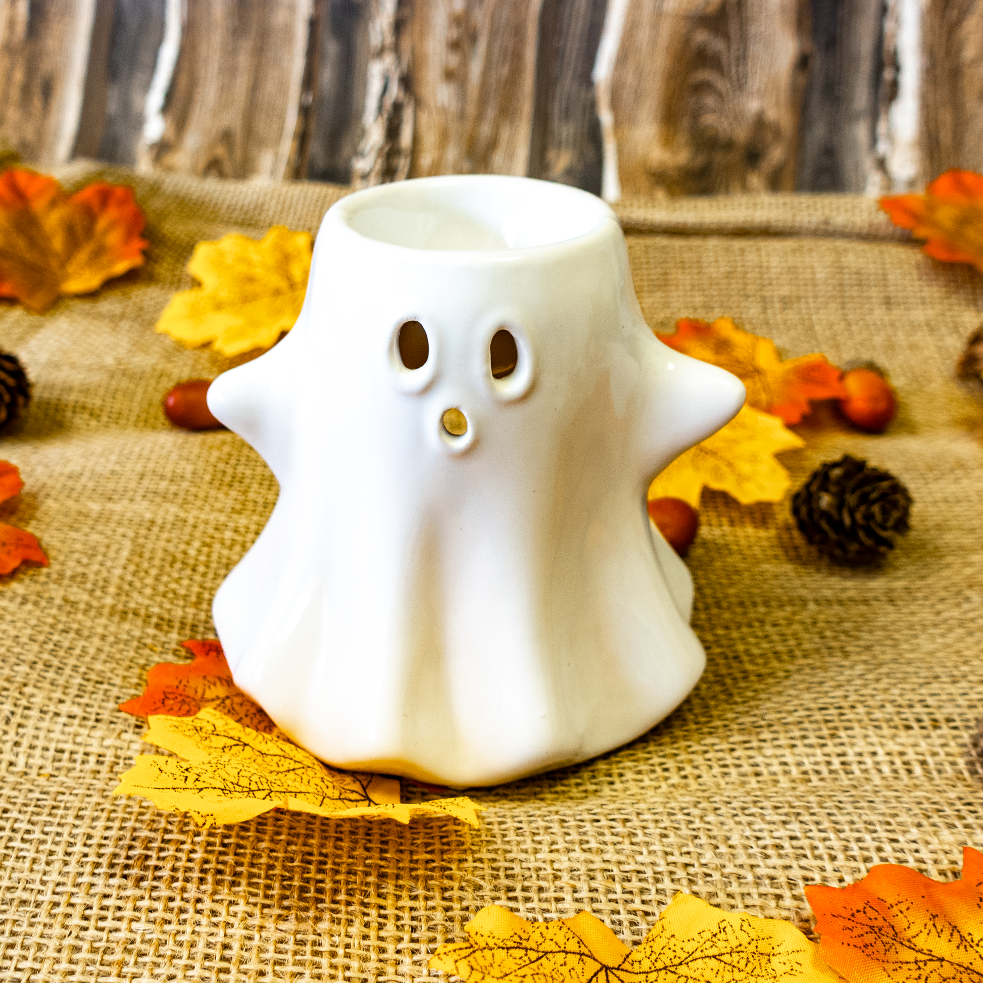 White ghost-shaped candle holder on a burlap surface with autumn leaves and pinecones.